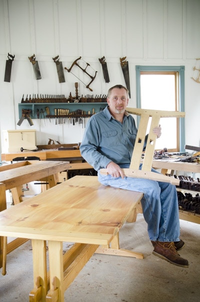 Will Myers Sitting On The Finished Trestle Table Holding A Trestle Table Leg Next To Woodworking Workbenches In Joshua Farnsworth'S Woodworking Workshop