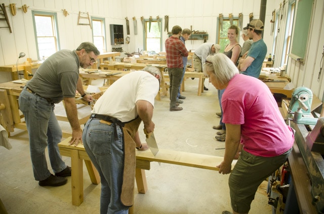 Woodworking Students Using An Antique Crosscut Panel Saw To Cut A Board With Woodworking Workbenches In The Background