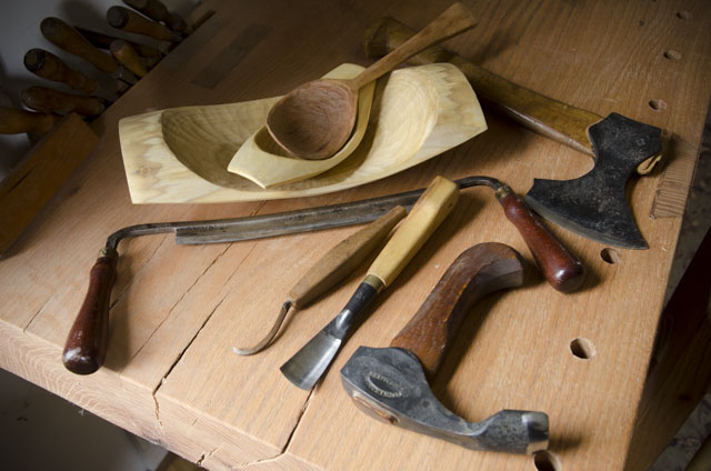 Green Woodworking Tools On A Woodworking Workbench, Including Carving Hatchet, Carving Gouge, Hook Knife, Bowl Adze, Draw Knife, And Wooden Spoon Inside A Wooden Bowl