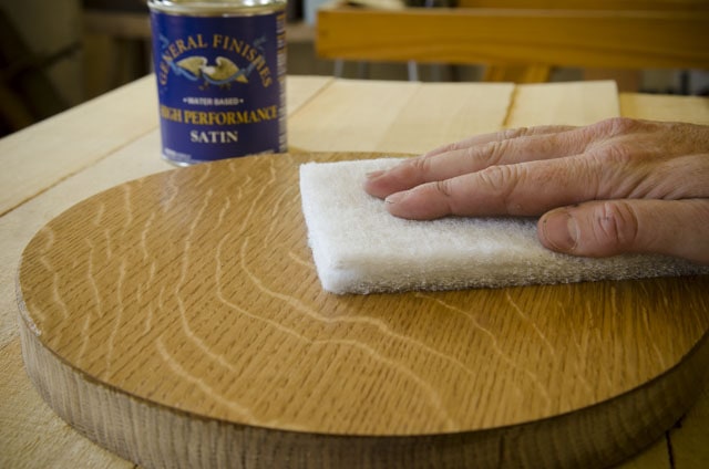 Man'S Hand Scuffing A Figured White Oak Piece Of Wood With A White 3M Scotch-Brite Abrasive Pad With A Can Of Water Based Wood Finish Made By General Finishes
