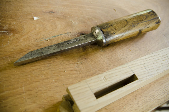 Best Chisel Guide: Pig Sticker Mortise Chisel Sitting Next To A Mortise On A Table Leg On A Woodworking Workbench